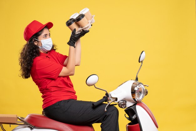 Corriere femminile vista frontale in maschera in bici con tazze di caffè su sfondo giallo servizio lavoratore lavoro pandemico consegna donna covid-