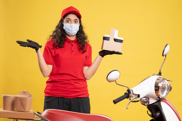 Corriere femminile vista frontale in uniforme rossa con tazze di caffè su sfondo giallo consegna del lavoratore covid- virus del servizio uniforme di lavoro pandemico