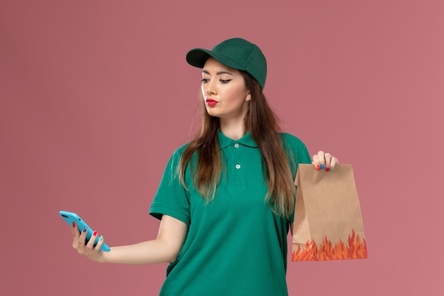 Corriere femminile vista frontale in uniforme verde utilizzando il telefono e tenendo il pacchetto di cibo sul lavoro di consegna uniforme servizio parete rosa