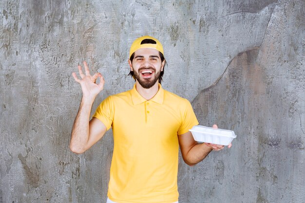 Corriere in uniforme gialla che consegna una scatola di plastica da asporto e mostra un segno positivo con la mano