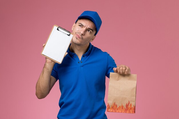 Corriere maschio di vista frontale in blocco note blu della tenuta uniforme che pensa sulla parete rosa, consegna di servizio uniforme del lavoratore