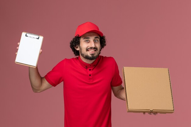 Corriere maschio di vista frontale in camicia rossa e mantello che tiene la scatola di cibo di consegna e il blocco note sulla parete rosa