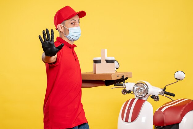 Corriere maschio di vista frontale in maschera con caffè di consegna e scatola sul lavoro di virus di colore pandemico uniforme di servizio giallo covid