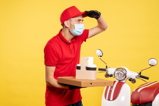 Corriere maschio di vista frontale in maschera con caffè di consegna e scatola sull'uniforme di lavoro di colore giallo del servizio covid-virus del lavoro