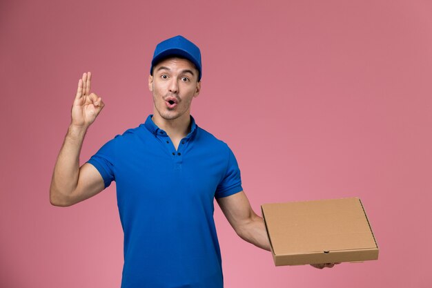 Corriere maschio di vista frontale in scatola di cibo della tenuta dell'uniforme blu che posa sulla parete rosa, consegna di servizio uniforme del lavoratore di lavoro