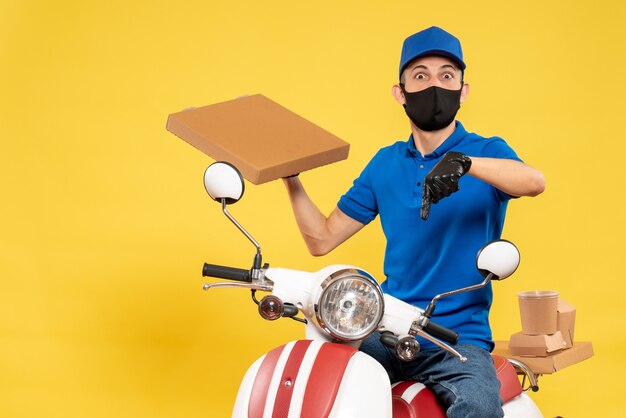 Corriere maschio di vista frontale in scatola di cibo della tenuta dell'uniforme blu sulla bici di lavoro di servizio di consegna di covid pandemia di virus giallo