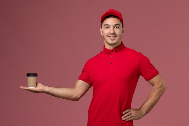 Corriere maschio di vista frontale in tazza di caffè di consegna uniforme rossa della tenuta sul lavoratore maschio della parete rosa