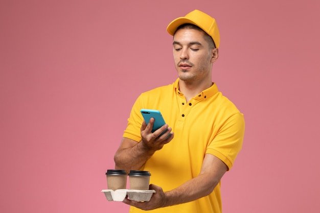 Corriere maschio di vista frontale in tazze di caffè di consegna della holding dell'uniforme gialla usando il suo telefono sulla scrivania rosa