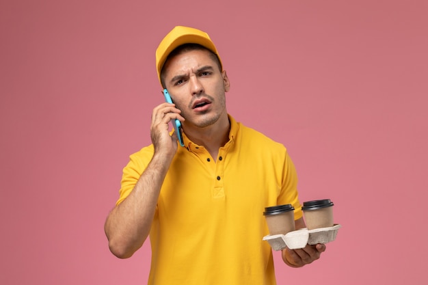 Corriere maschio di vista frontale in tazze di caffè di consegna della tenuta della tenuta uniforme gialla che comunicano sul telefono sui precedenti rosa