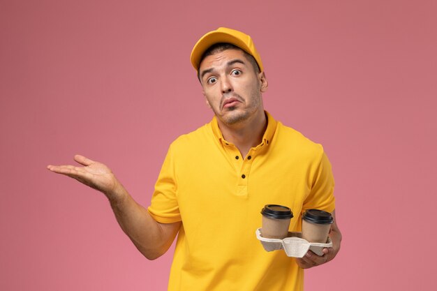 Corriere maschio di vista frontale in tazze di caffè di consegna della tenuta uniforme gialla sui precedenti rosa chiaro