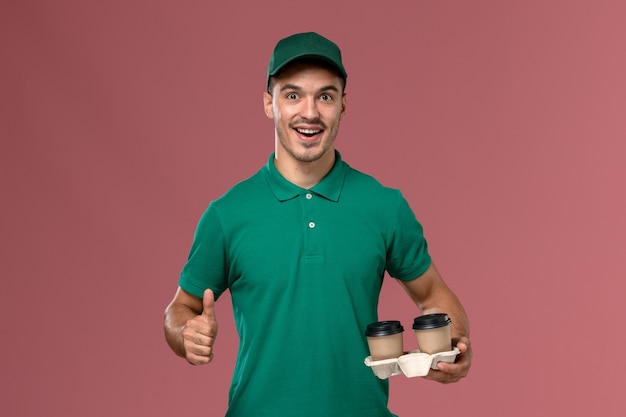 Corriere maschio di vista frontale in tazze di caffè di consegna della tenuta uniforme verde con il sorriso su fondo rosa