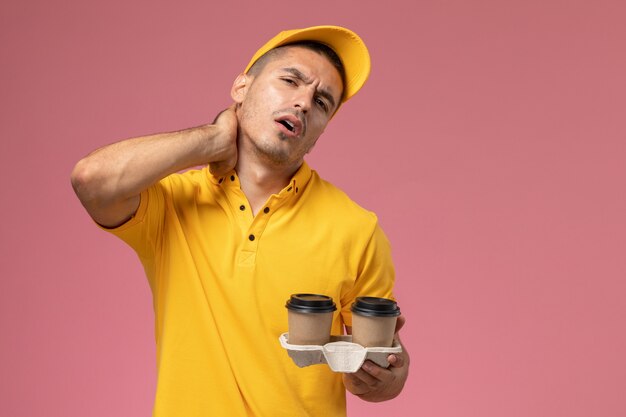 Corriere maschio di vista frontale in tazze di caffè di consegna uniformi gialle della holding che hanno neckache su fondo rosa