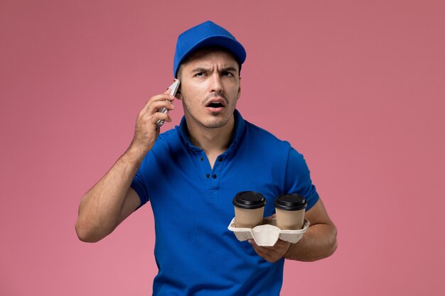 Corriere maschio di vista frontale in uniforme blu che parla sul telefono e che tiene le tazze di caffè sulla parete rosa, consegna di lavoro di servizio uniforme