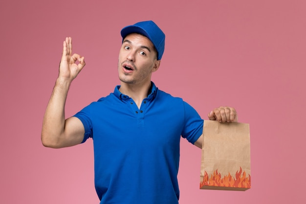 Corriere maschio di vista frontale in uniforme blu che tiene il pacchetto alimentare di carta sulla parete rosa, consegna del lavoro di servizio uniforme del lavoratore