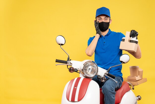 Corriere maschio di vista frontale in uniforme blu con caffè sulla bici pandemica gialla covid - servizio di lavoro virus consegna lavoro