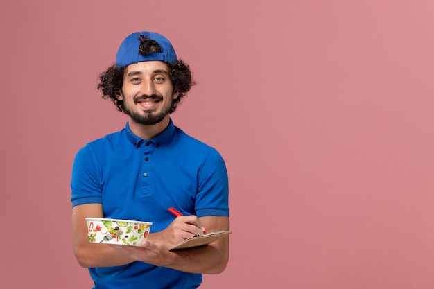 Corriere maschio di vista frontale in uniforme blu e mantello che tiene blocco note e ciotola di consegna rotonda che scrive sulla parete rosa