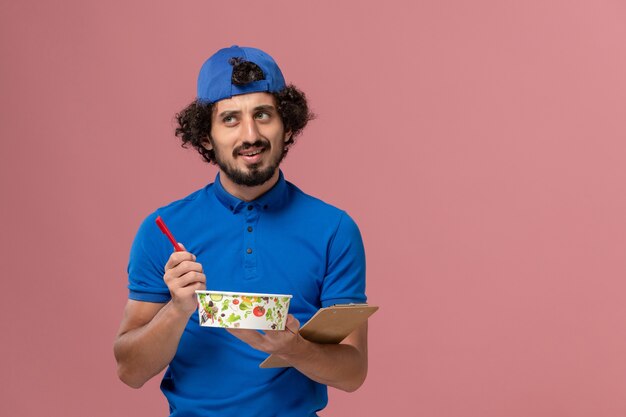 Corriere maschio di vista frontale in uniforme blu e mantello che tiene blocco note e ciotola di consegna rotonda che scrive sulla parete rosa