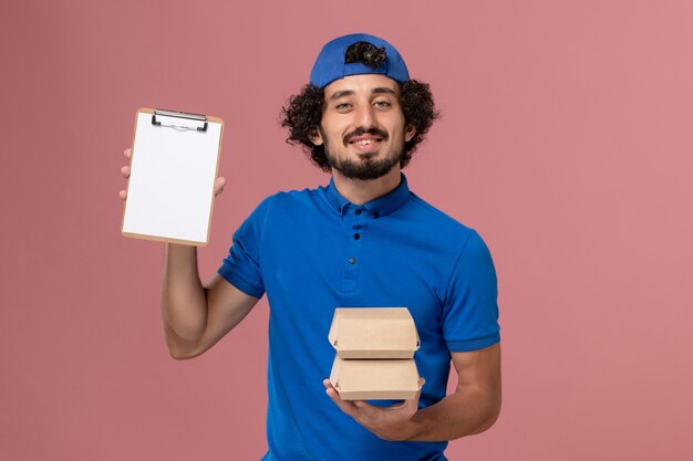 Corriere maschio di vista frontale in uniforme blu e mantello che tiene i pacchetti di cibo di consegna con il blocco note sul servizio di consegna del lavoro uniforme della parete rosa