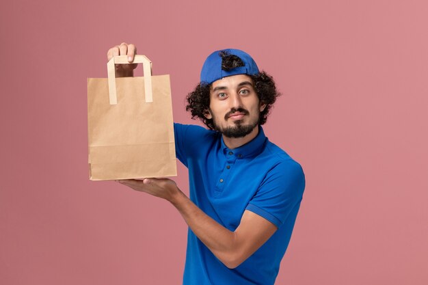 Corriere maschio di vista frontale in uniforme blu e mantello che tiene il pacchetto di cibo di carta di consegna sul maschio dell'uniforme di servizio di consegna della parete rosa