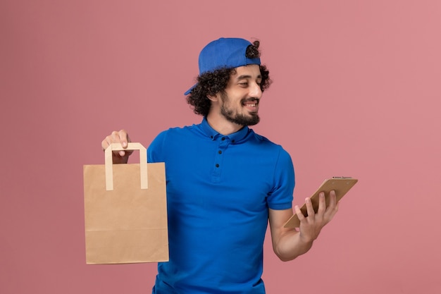 Corriere maschio di vista frontale in uniforme blu e mantello che tiene il pacchetto di cibo di consegna e blocco note sull'uniforme di servizio di lavoro di consegna della parete rosa