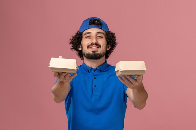 Corriere maschio di vista frontale in uniforme blu e mantello che tiene piccoli pacchetti di cibo di consegna sulla parete rosa