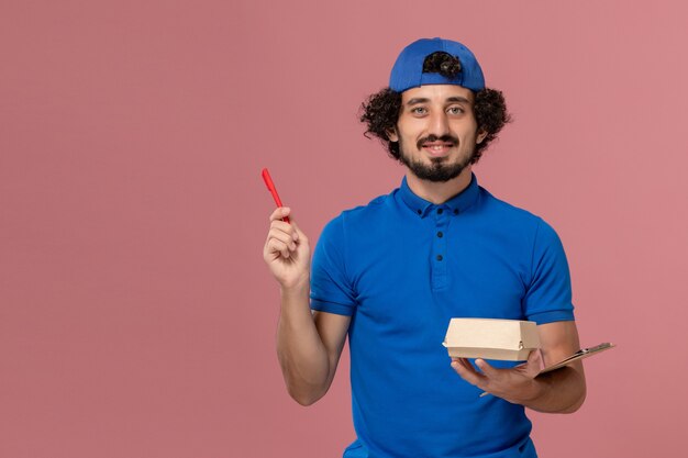 Corriere maschio di vista frontale in uniforme blu e mantello che tiene piccolo pacchetto di cibo di consegna e blocco note che scrive sul servizio di consegna del lavoro uniforme della parete rosa