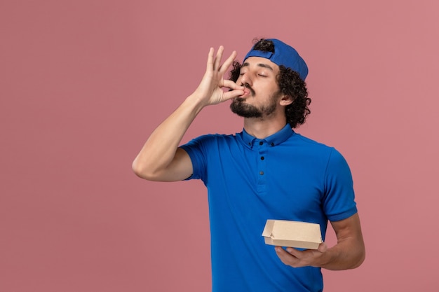 Corriere maschio di vista frontale in uniforme blu e mantello che tiene piccolo pacchetto di cibo di consegna sulla parete rosa