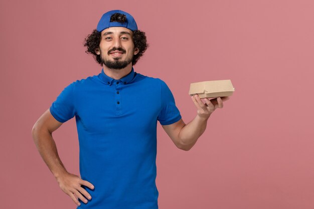 Corriere maschio di vista frontale in uniforme blu e mantello che tiene piccolo pacchetto di cibo di consegna sulla parete rosa