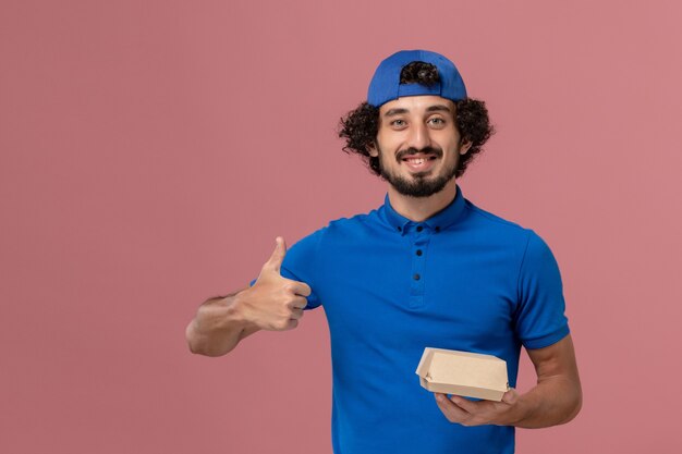 Corriere maschio di vista frontale in uniforme blu e mantello che tiene piccolo pacchetto di cibo di consegna sulla parete rosa