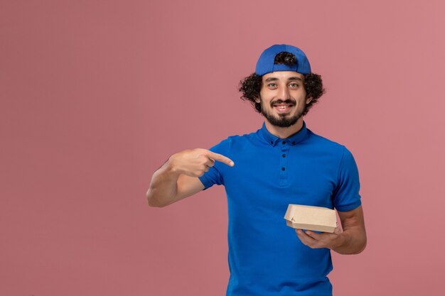 Corriere maschio di vista frontale in uniforme blu e mantello che tiene piccolo pacchetto di cibo di consegna sulla parete rosa