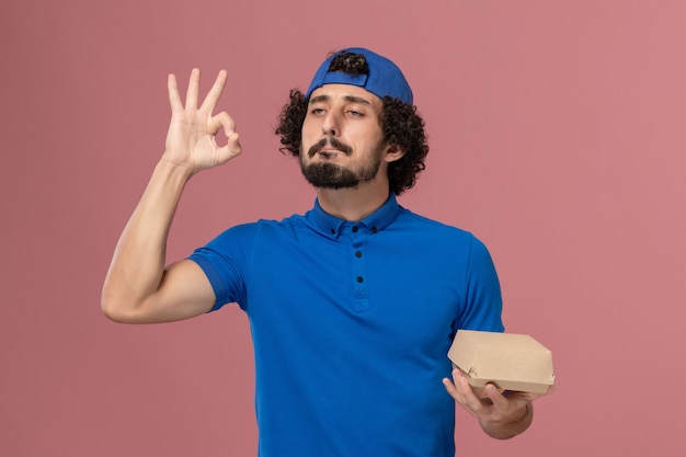 Corriere maschio di vista frontale in uniforme blu e mantello che tiene piccolo pacchetto di cibo di consegna sulla parete rosa