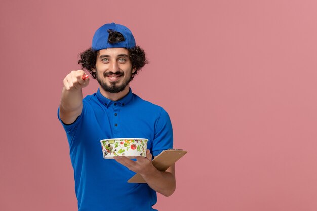 Corriere maschio di vista frontale in uniforme blu e penna del blocco note della tenuta del capo e ciotola rotonda di consegna sulla parete rosa