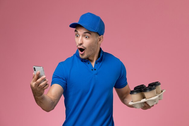 Corriere maschio di vista frontale in uniforme blu facendo uso delle tazze di caffè della tenuta del telefono sulla parete rosa, consegna di servizio uniforme del lavoratore