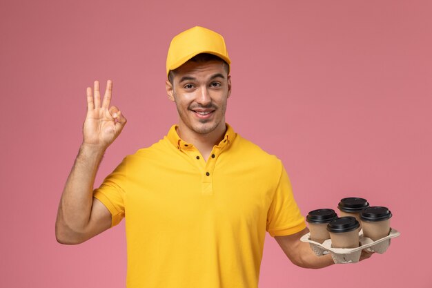 Corriere maschio di vista frontale in uniforme gialla che sorride e che tiene le tazze di caffè di consegna sullo scrittorio rosa