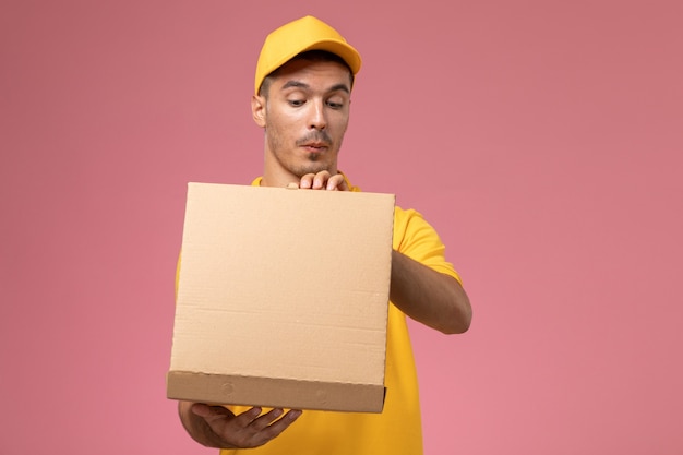 Corriere maschio di vista frontale in uniforme gialla che tiene e che apre la scatola di consegna del cibo sullo scrittorio rosa
