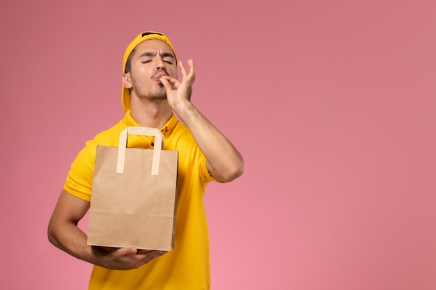 Corriere maschio di vista frontale in uniforme gialla che tiene il pacchetto alimentare di consegna della carta sui precedenti rosa.