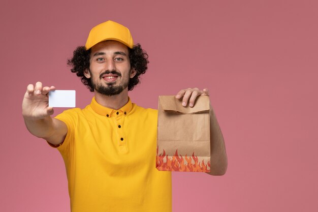 Corriere maschio di vista frontale in uniforme gialla che tiene il pacchetto di cibo e la carta di plastica sulla parete rosa