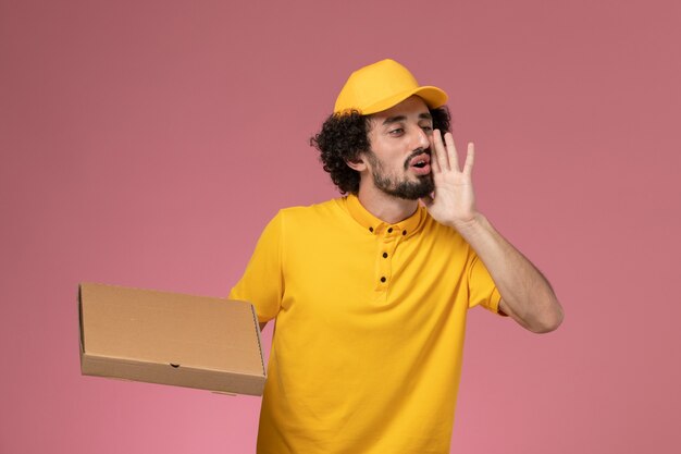 Corriere maschio di vista frontale in uniforme gialla che tiene la scatola di consegna del cibo che chiama sul muro rosa chiaro