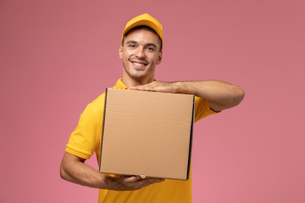 Corriere maschio di vista frontale in uniforme gialla che tiene la scatola di consegna del cibo e che sorride sui precedenti rosa