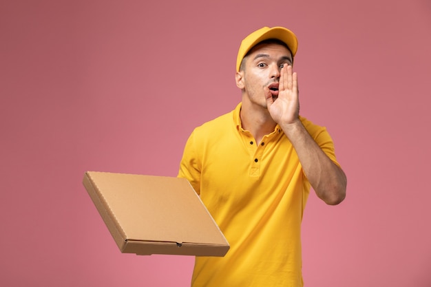 Corriere maschio di vista frontale in uniforme gialla che tiene la scatola di consegna del cibo e chiama qualcuno sullo sfondo rosa