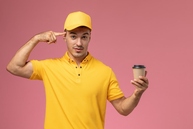 Corriere maschio di vista frontale in uniforme gialla che tiene la tazza di consegna del caffè marrone che tocca la sua tempia sulla scrivania rosa