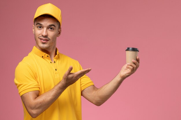 Corriere maschio di vista frontale in uniforme gialla che tiene tazza di consegna del caffè sui precedenti rosa