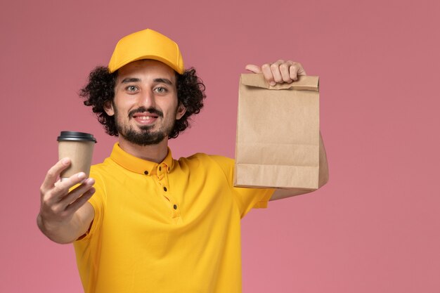 Corriere maschio di vista frontale in uniforme gialla e mantello che tiene la tazza di caffè di consegna e il pacchetto di cibo sulla parete rosa