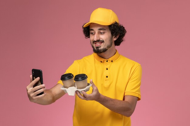 Corriere maschio di vista frontale in uniforme gialla e mantello che tiene le tazze di caffè marroni di consegna che prendono la foto sulla parete rosa