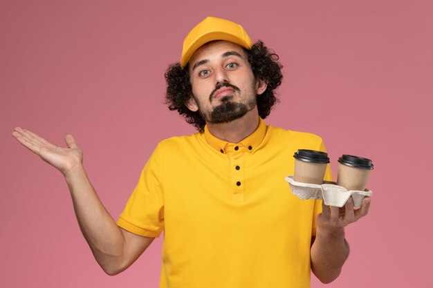 Corriere maschio di vista frontale in uniforme gialla e mantello che tiene le tazze di caffè marroni di consegna sulla parete rosa