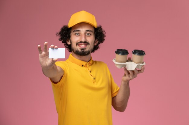 Corriere maschio di vista frontale in uniforme gialla e mantello che tiene le tazze e la carta di caffè di consegna marroni sulla parete rosa-chiaro