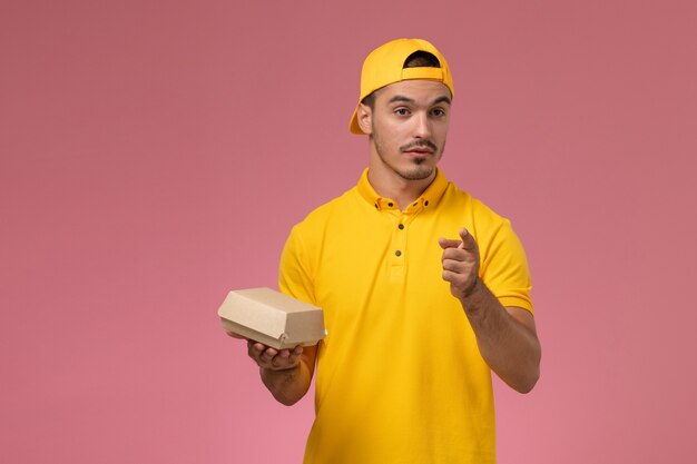 Corriere maschio di vista frontale in uniforme gialla e mantello che tiene piccolo pacchetto di cibo di consegna e parlando su sfondo rosa chiaro.