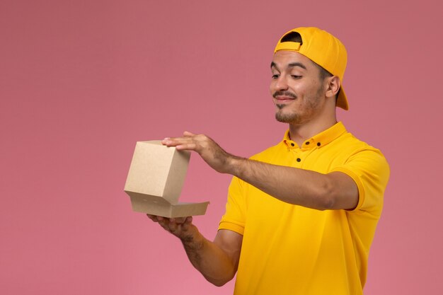 Corriere maschio di vista frontale in uniforme gialla e mantello che tiene piccolo pacchetto di cibo di consegna su fondo rosa.