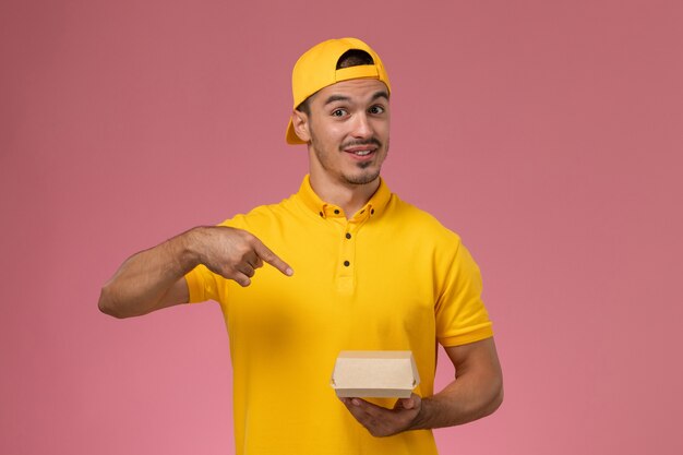 Corriere maschio di vista frontale in uniforme gialla e mantello che tiene piccolo pacchetto di cibo di consegna su fondo rosa.