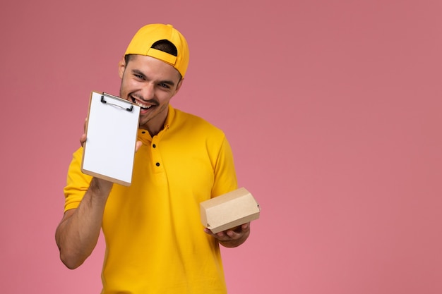 Corriere maschio di vista frontale in uniforme gialla e mantello che tiene poco pacchetto di cibo di consegna e blocco note sulla scrivania rosa.